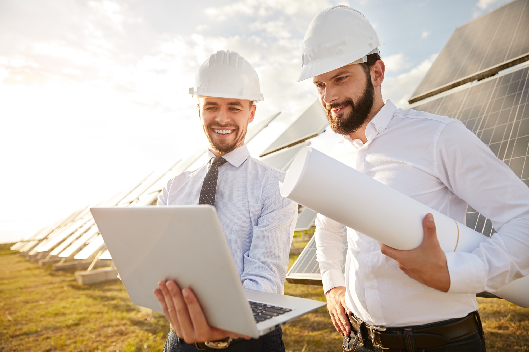 Smiling Professional Engineer With Laptop Discussing Details of Project With Colleague While Standing Near Solar Panels for Alternative Energy in Field.